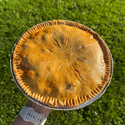 A freshly baked golden-brown chicken pie held up by a person's hand against a bright green grassy background.